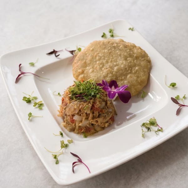 A white rectangular plate holds a savory crab cake and a golden fried flatbread, garnished with microgreens and an orchid.
