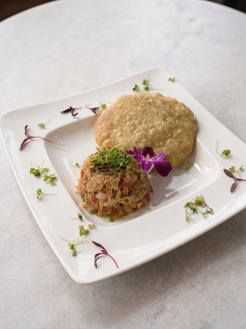 A white rectangular plate holds a savory crab cake and a golden fried flatbread, garnished with microgreens and an orchid.