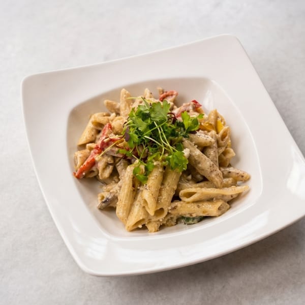 Creamy penne pasta with red peppers and fresh herbs in a white square bowl on a light table.